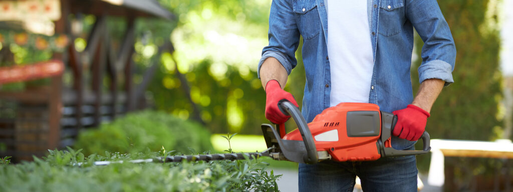 Anonymous muscular handyman in protective gloves using electric trimmer for shaping bushes on backyard. Crop view of male worker cutting hedge, while doing seasonal work outdoor. Concept of gardening.